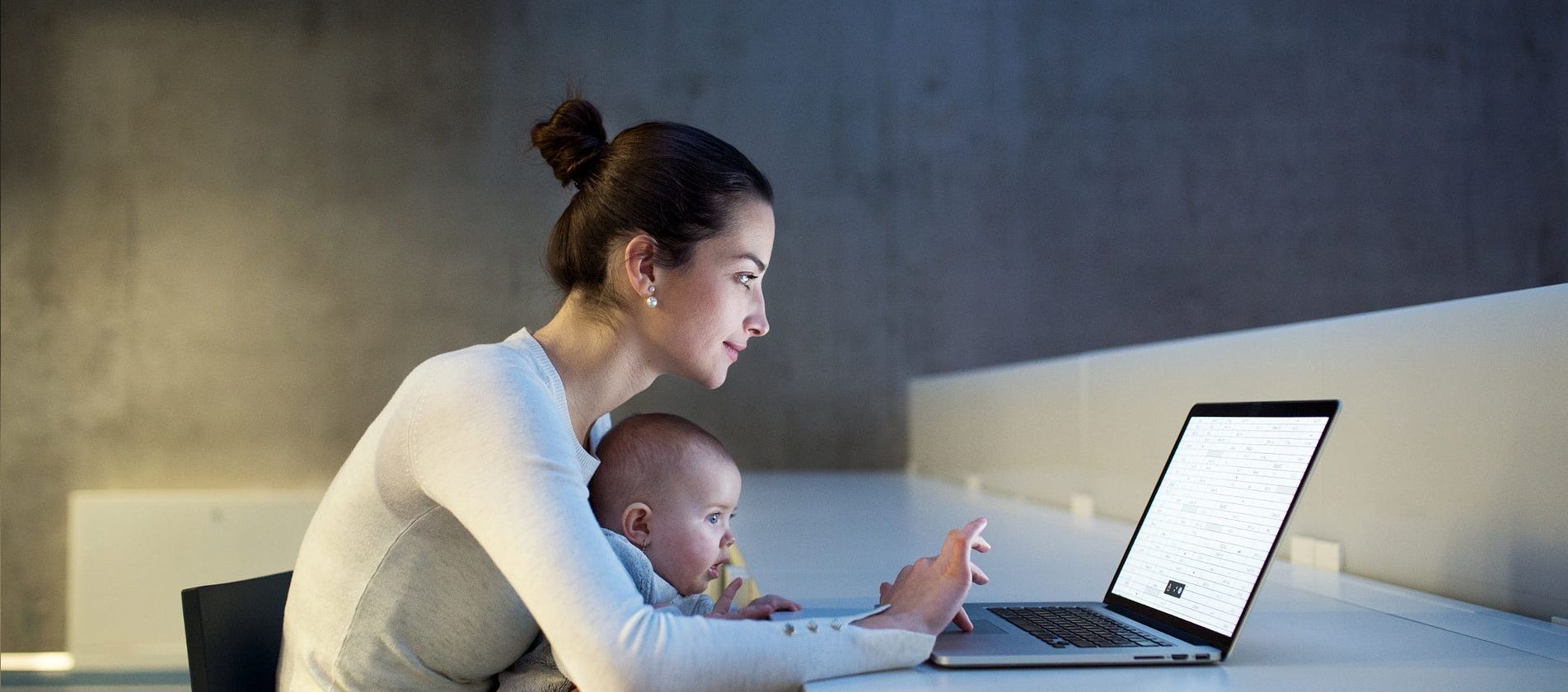 Young student with a baby sitting on desk in room in a library or office, using laptop.